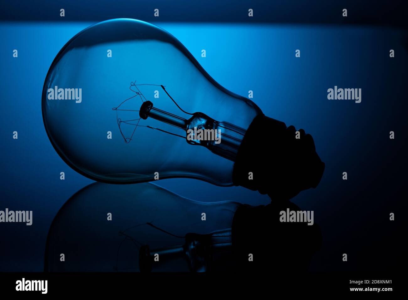 An incandescent light bulb lies on reflective glass, against a blue background, in a dark key. Stock Photo