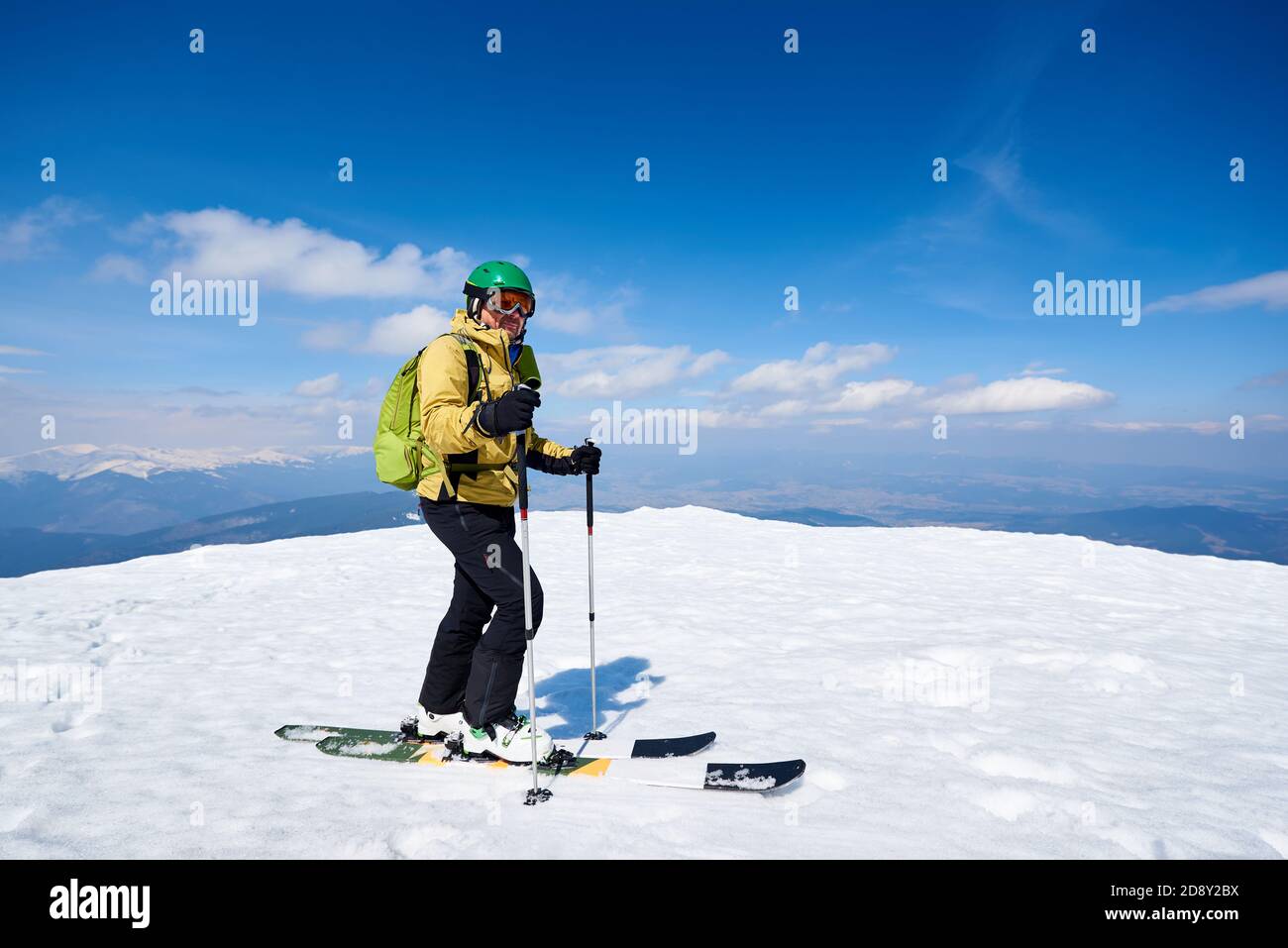 Sportsman skier with backpack in helmet and goggles standing on skis on snowy mountain summit on copy space background of bright blue sky and highland landscape. Professional skiing equipment concept. Stock Photo