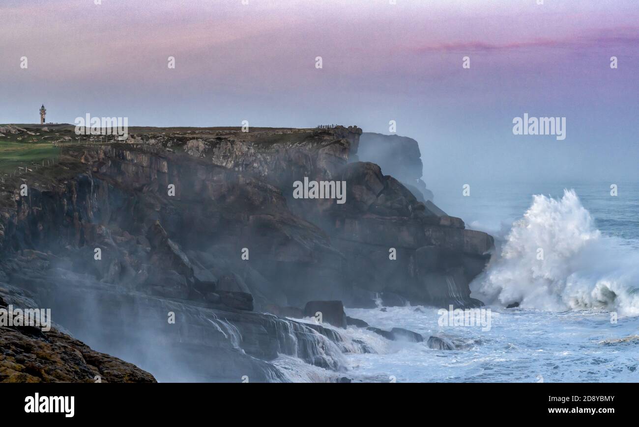 huge storm waves crash onto the Cape Ajo in Spain with the lighthouse on the cliffs above at sunrise Stock Photo