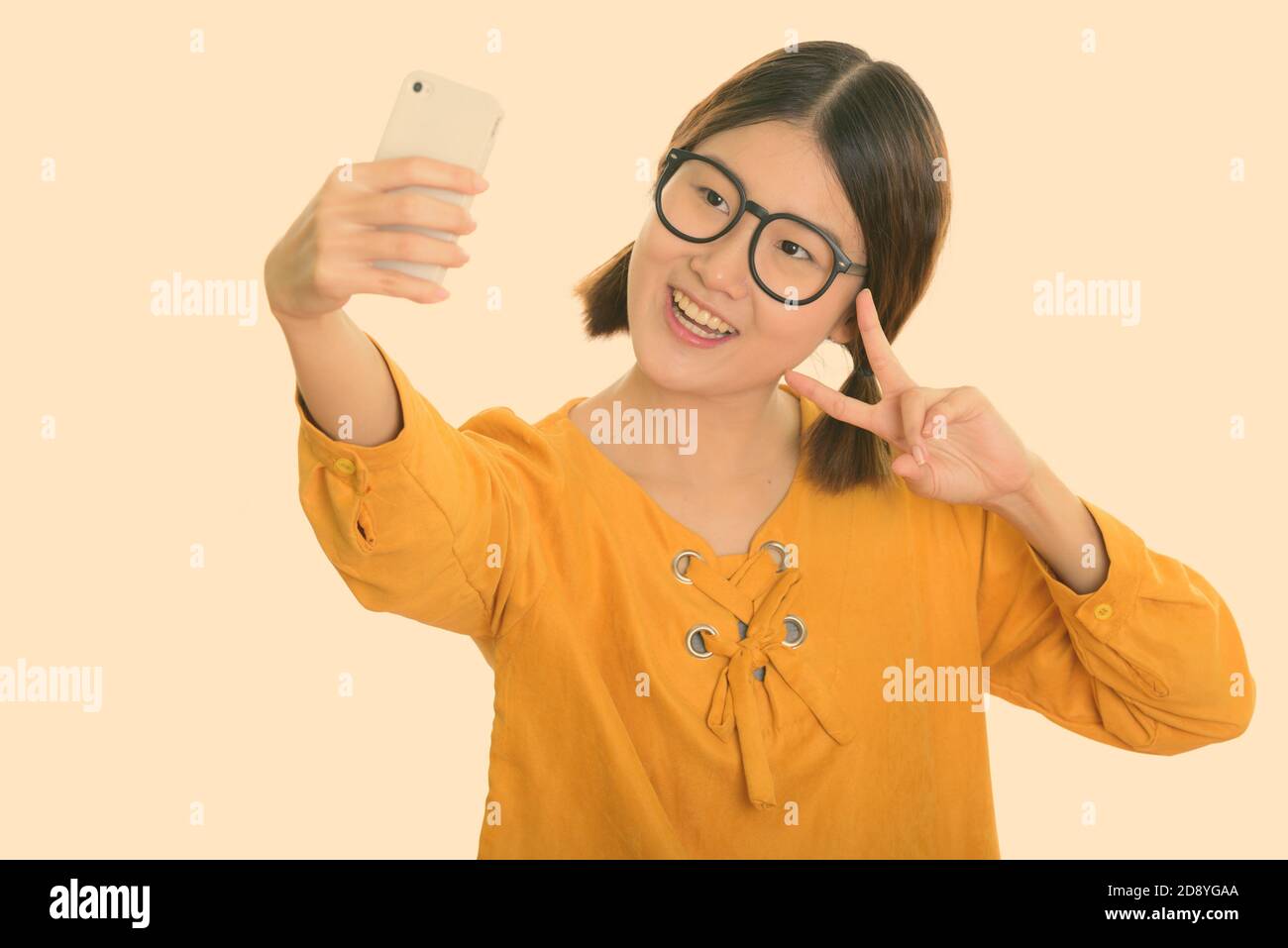 Young happy Asian woman smiling while taking selfie picture with mobile phone and giving peace sign Stock Photo