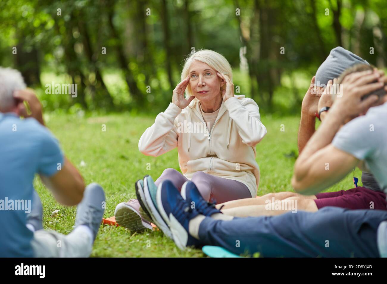 Group of modern senior people gathered together in park sitting on mats on grass doing neck stretching relaxing exercise Stock Photo