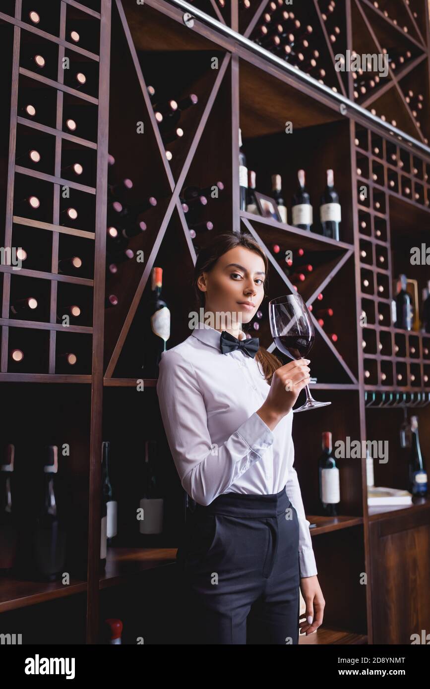 Young sommelier with glass of wine looking at camera near racks with ...