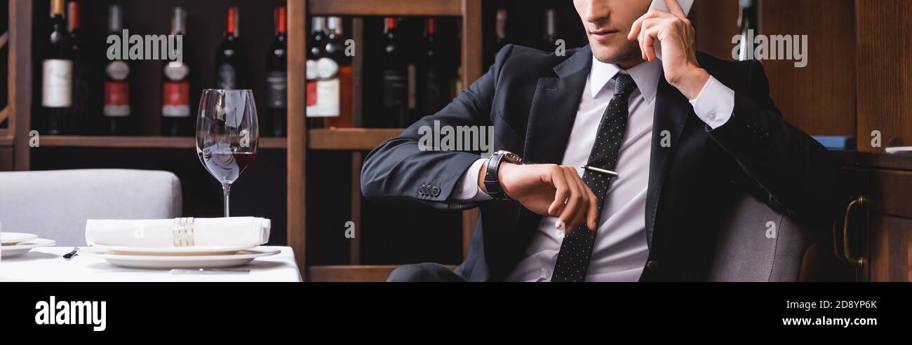 Panoramic shot of man in suit talking on smartphone and checking time on wristwatch near glass of wine in restaurant Stock Photo