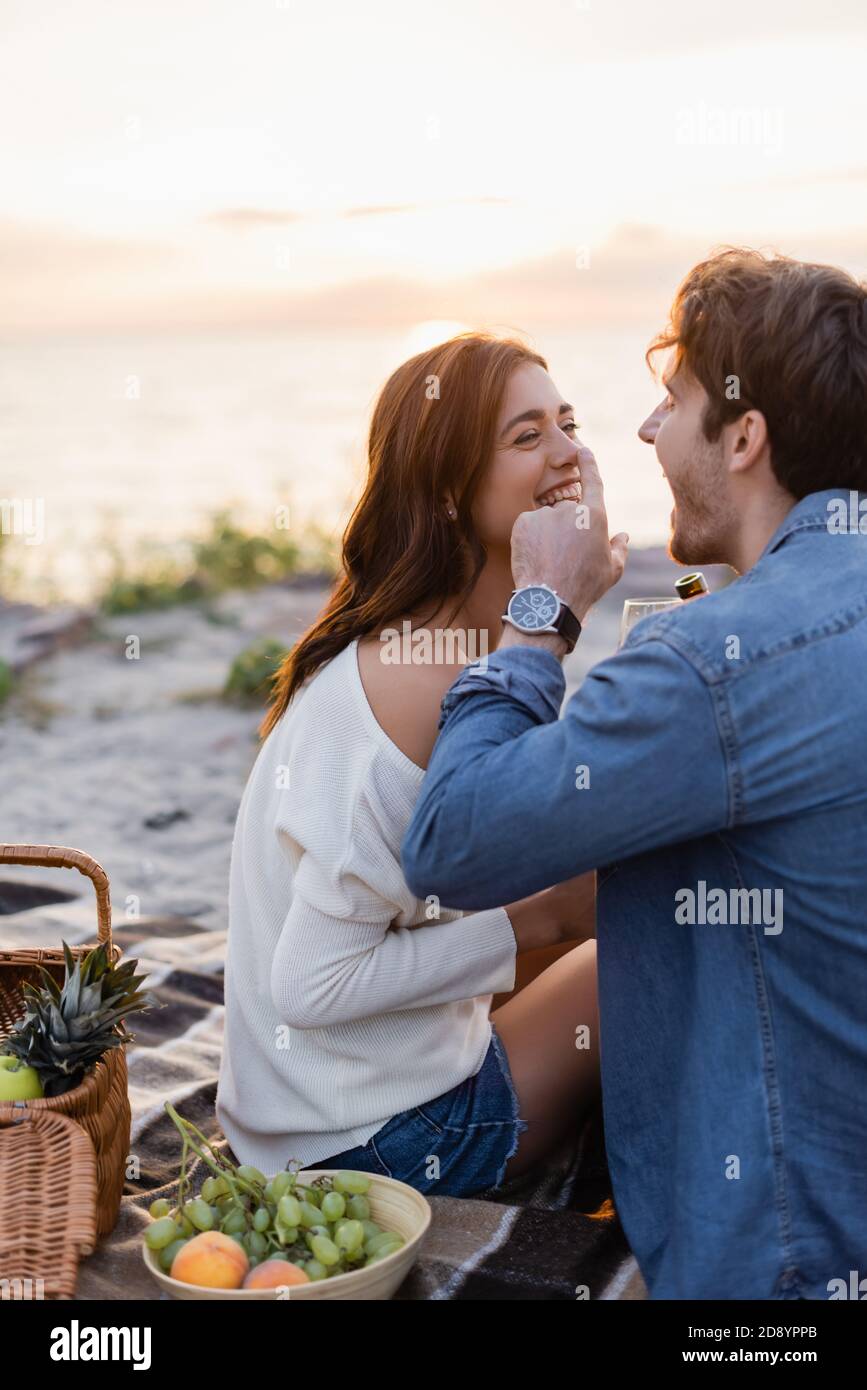 Selective focus of man with bottle of wine laughing while touching nose of girlfriend during picnic on beach Stock Photo