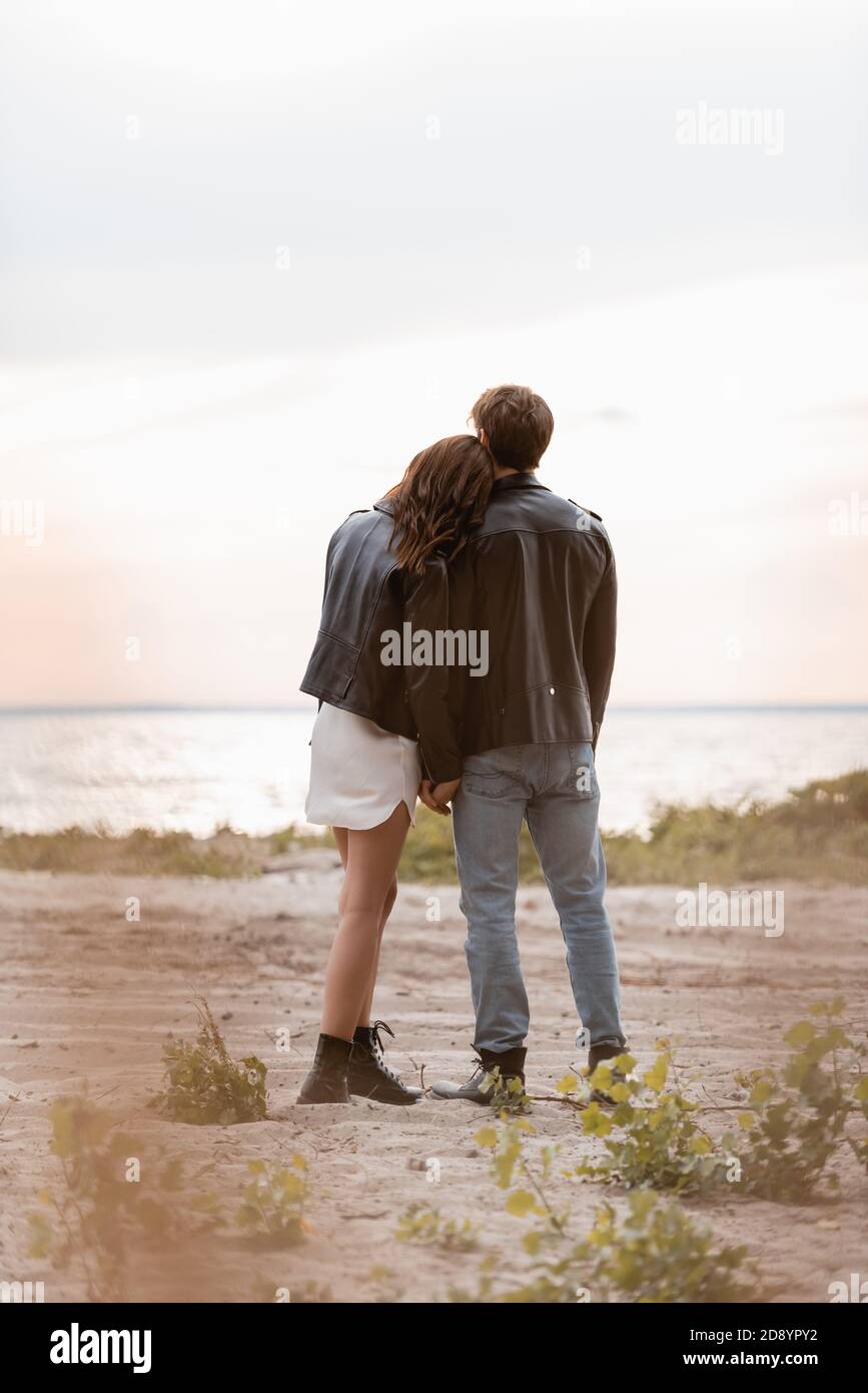 Back view of couple in leather jackets standing on sea beach at sunset ...