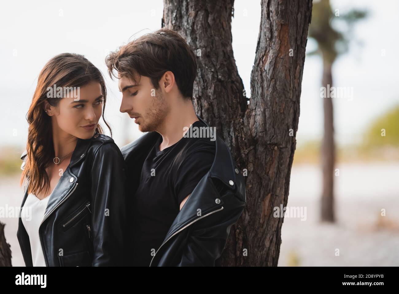 Brunette woman looking at boyfriend in leather jacket near tree in ...