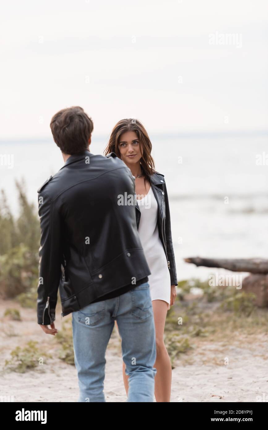 Selective focus of young woman in dress and leather jacket looking at ...