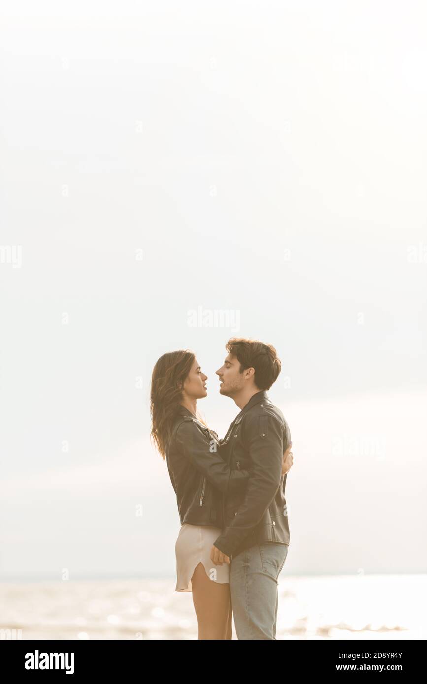 Side view of woman in leather jacket hugging boyfriend with sea and sky ...