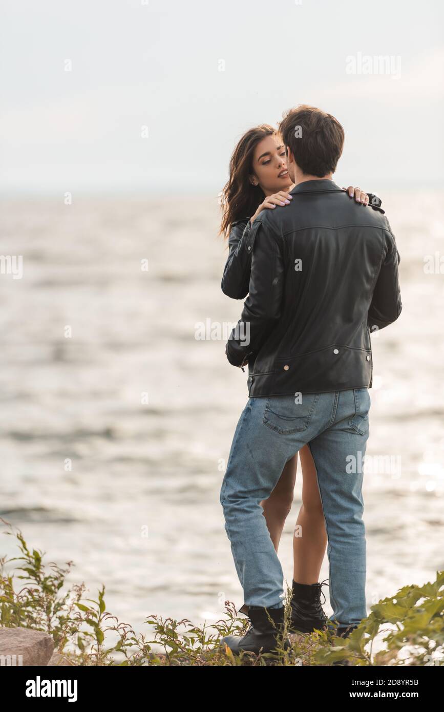 Brunette woman embracing boyfriend in leather jacket and jeans on beach ...