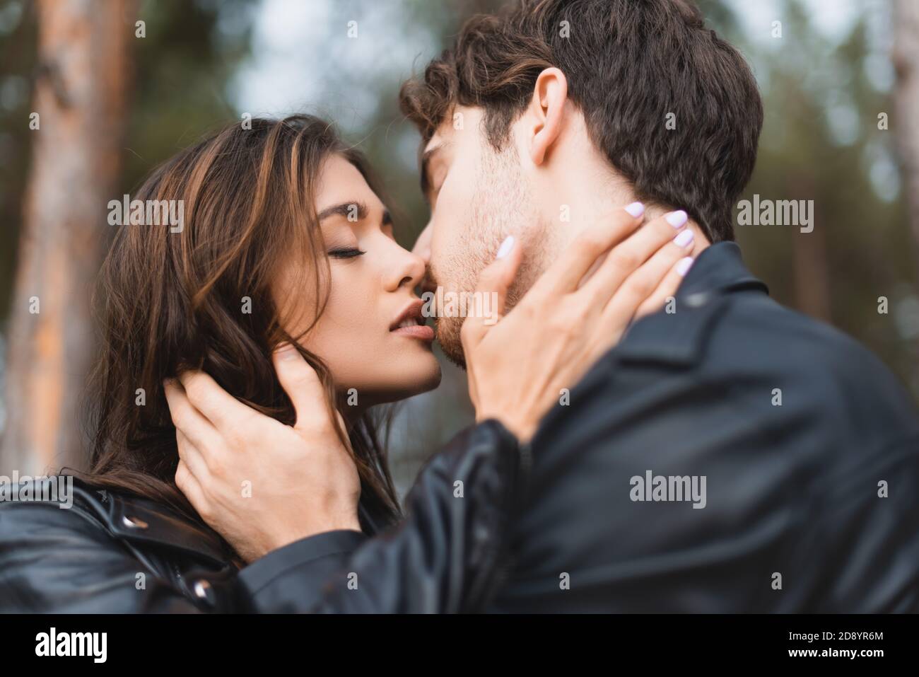 Selective focus of young couple in leather jackets kissing in forest ...