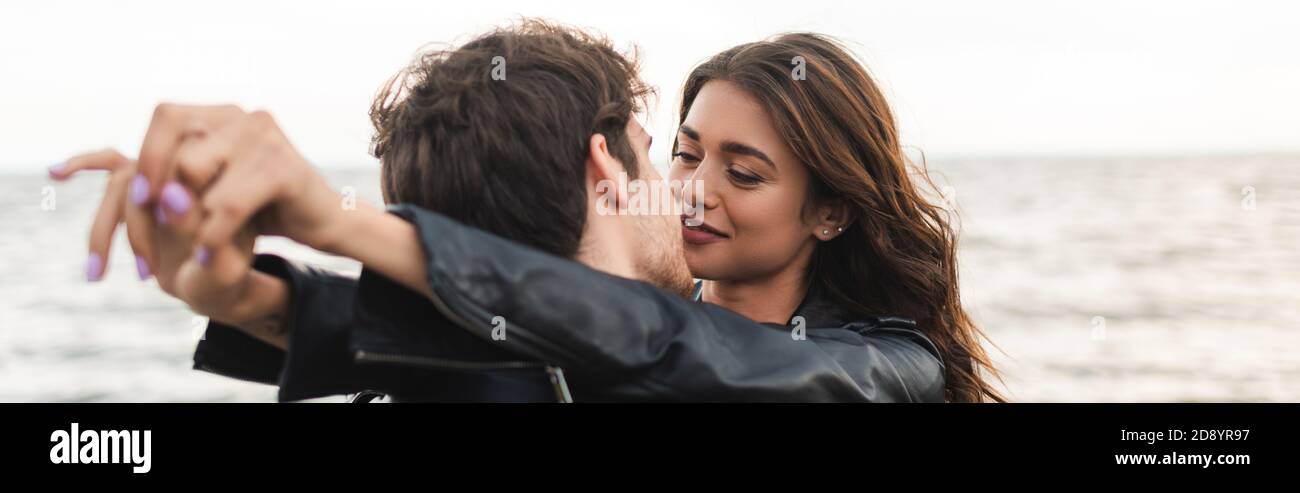 Panoramic shot of man kissing girlfriend in leather jacket near sea ...