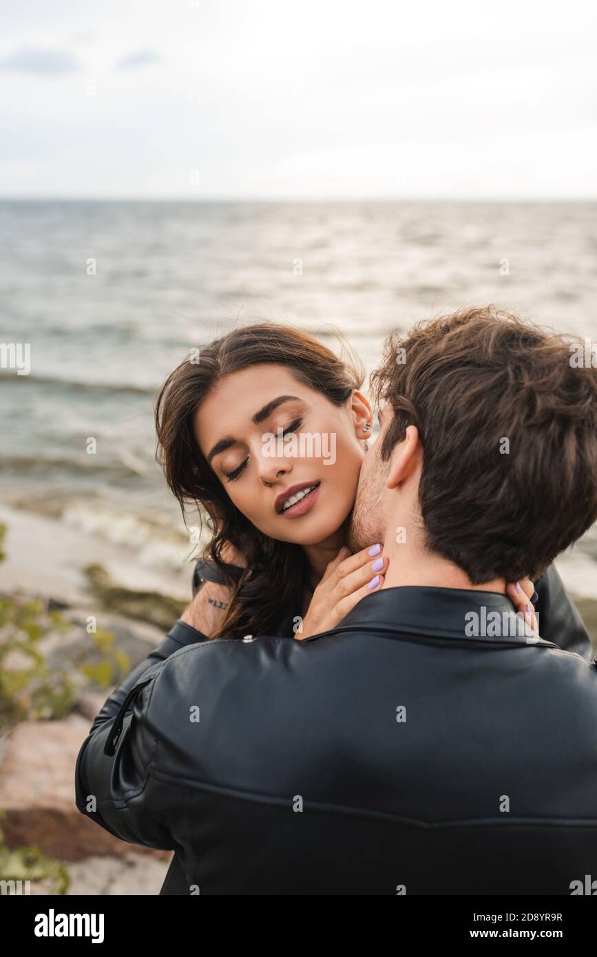 Man in black leather jacket kissing girlfriend in neck near sea on ...