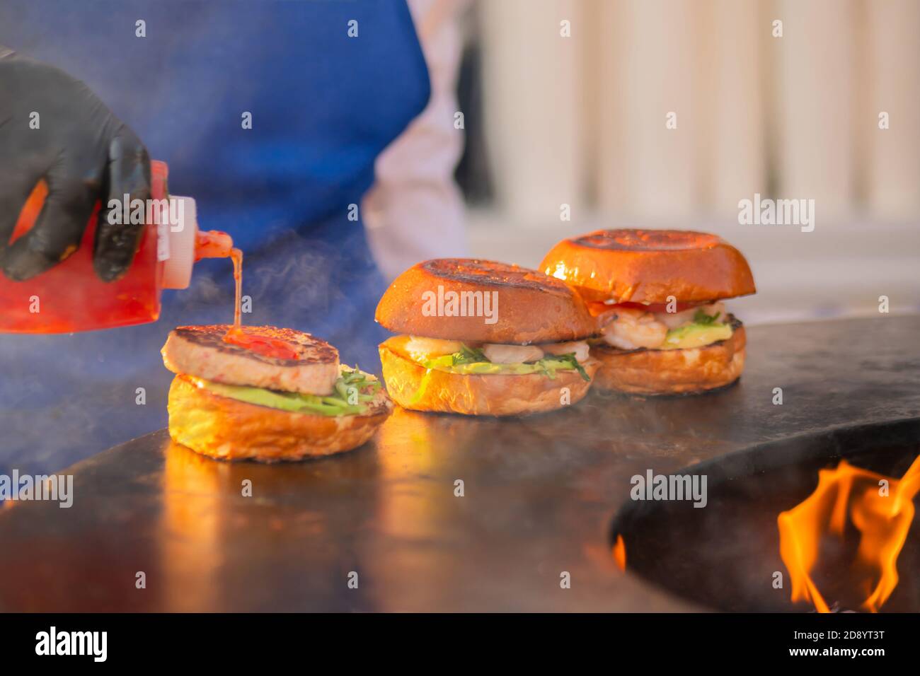 Chef preparing burgers with seafood, fish cutlet, shrimp, prawn on brazier with hot flame at summer local food market - close up view. Outdoor cooking Stock Photo