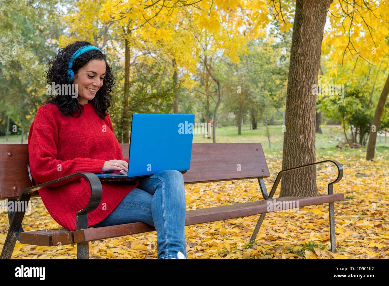 young woman with curly hair sitting on a bench working with laptop and blue wireless headphones in autumn with leaves falling from trees Stock Photo