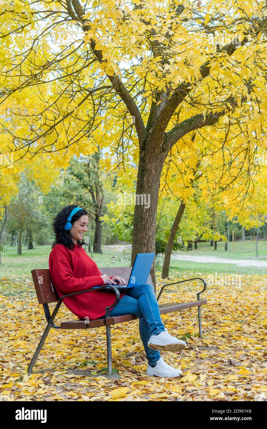 young woman sitting on a bench working with laptop and blue wireless headphones in autumn with leaves falling from trees Stock Photo