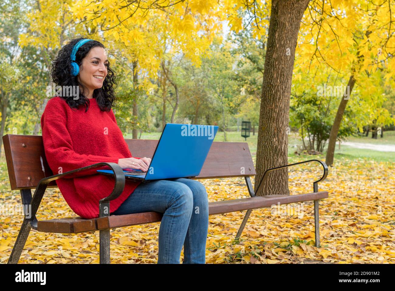 young woman with curly hair sitting on a bench working with laptop and blue wireless headphones while smiling in autumn with leaves falling from the t Stock Photo