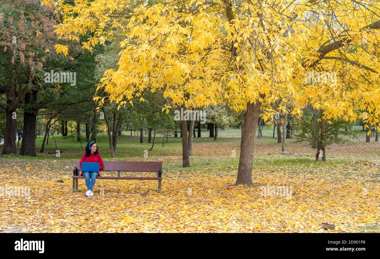 young brunette woman sitting in a park in autumn with her laptop and blue headphones. background of colored trees and fallen leaves Stock Photo
