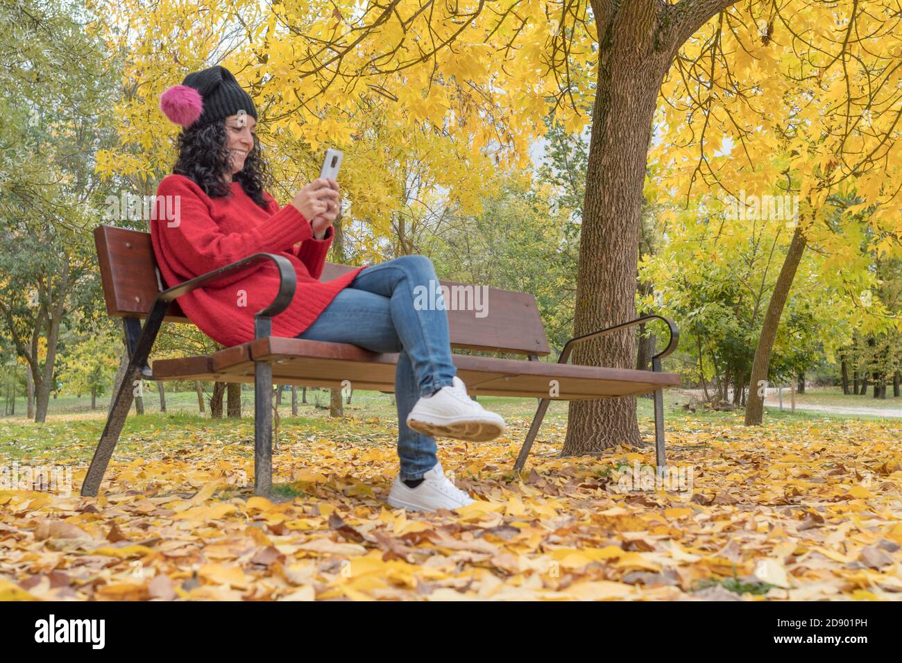 young woman in a woolen hat sitting on a wooden bench in a park laughing while chatting with her mobile. background of colored trees and fallen leaves Stock Photo