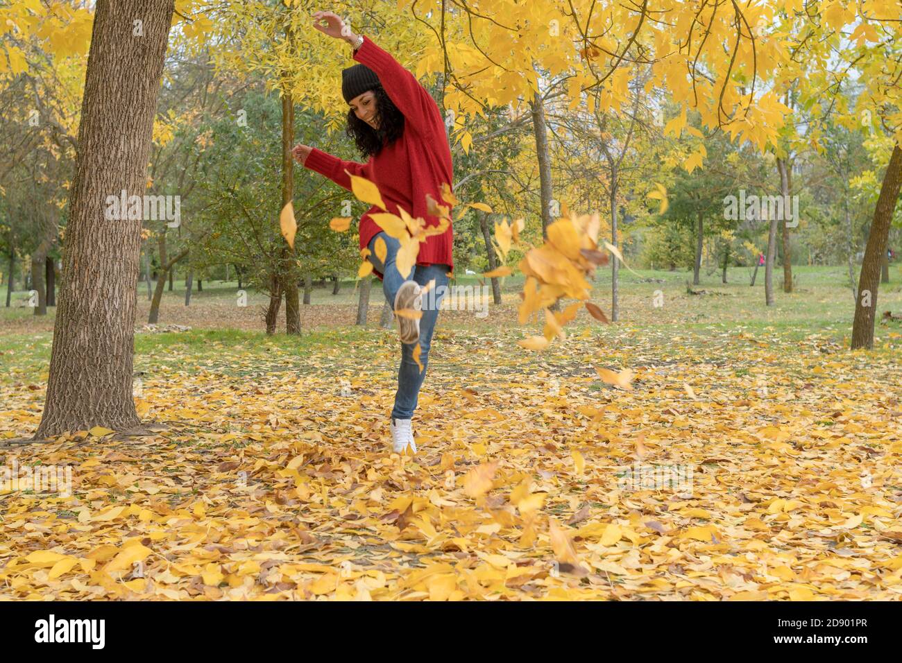 young woman with curly hair and hat and wool sweater has fun kicking a pile of dry leaves that have fallen from the trees in a park in autumn Stock Photo