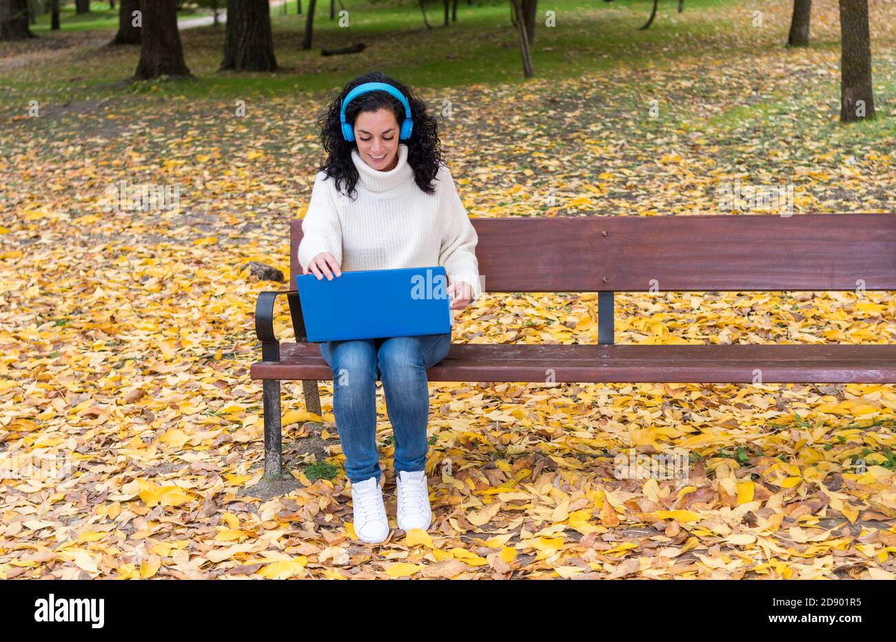 young woman with curly hair sitting on a bench working with laptop and blue wireless headphones while smiling in autumn with leaves falling from the t Stock Photo