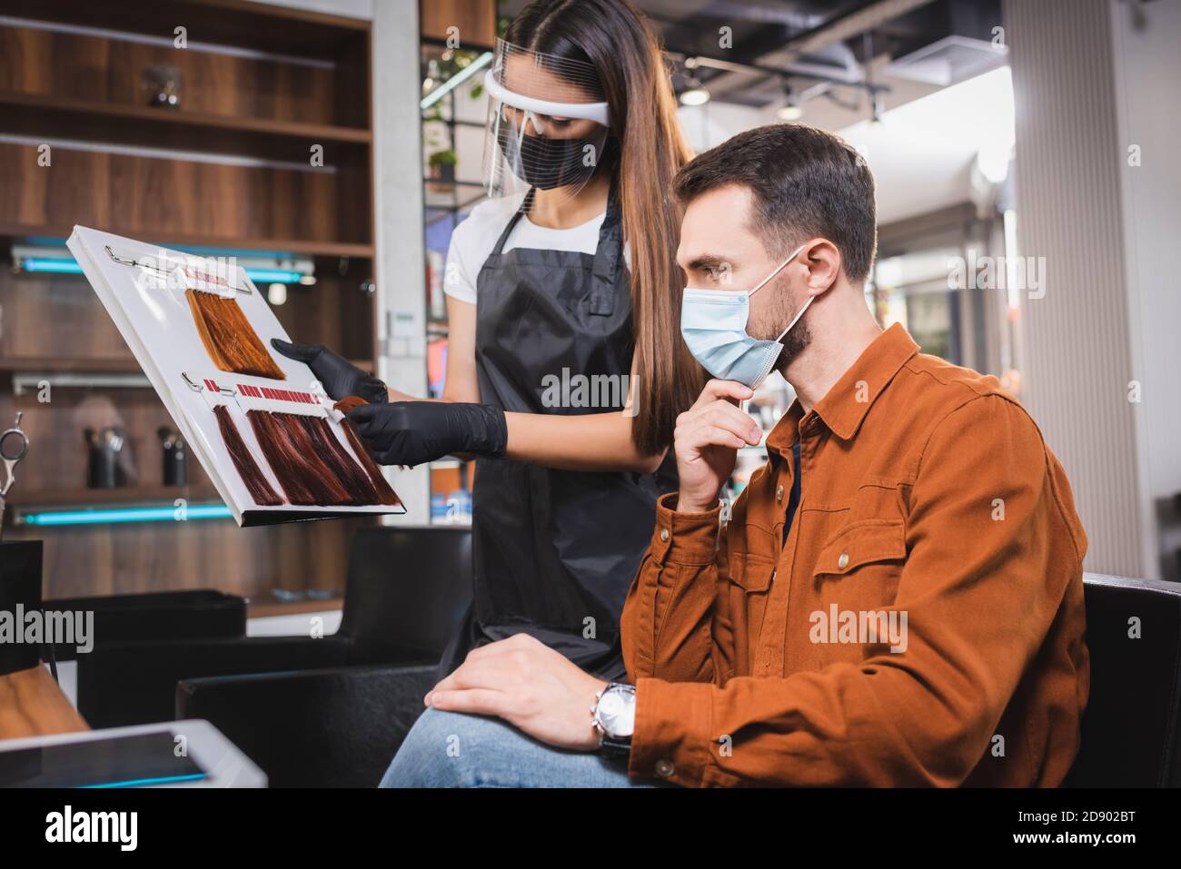 hairstylist in face shield and apron showing hair colors palette to man in medical mask Stock Photo