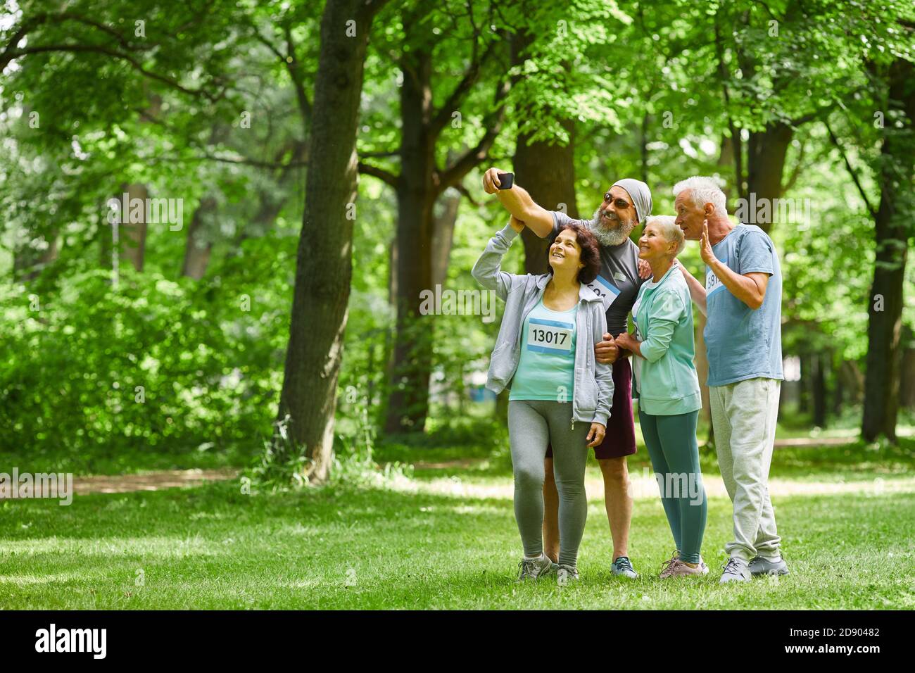 Group of joyful sporty senior adult men and women standing together in park taking group selfie shot on smart phone camera Stock Photo