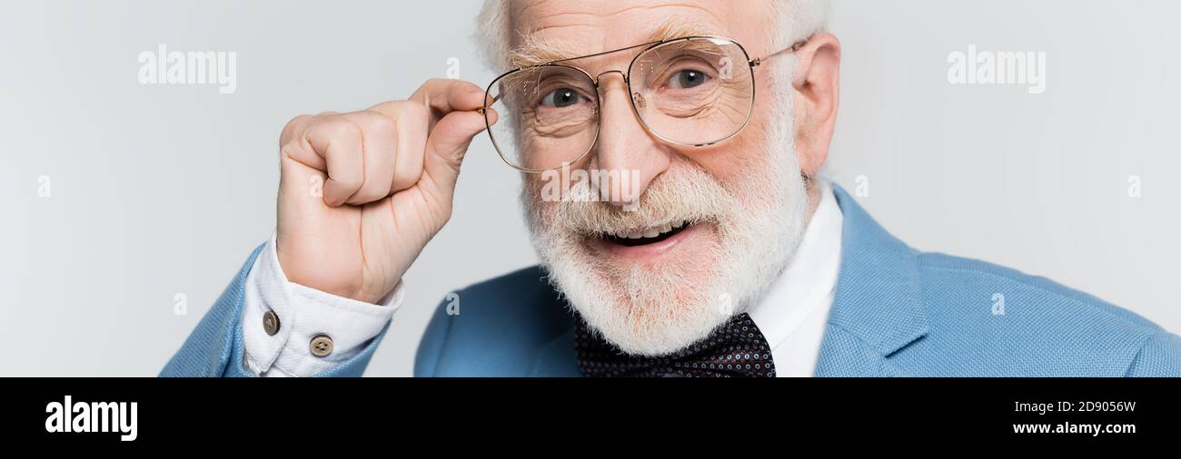 Smiling elderly man touching eyeglasses isolated on grey, banner Stock Photo