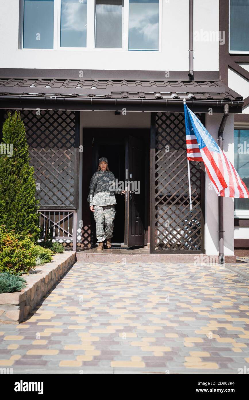 Military servicewoman standing in doorway near american flag and bushes Stock Photo
