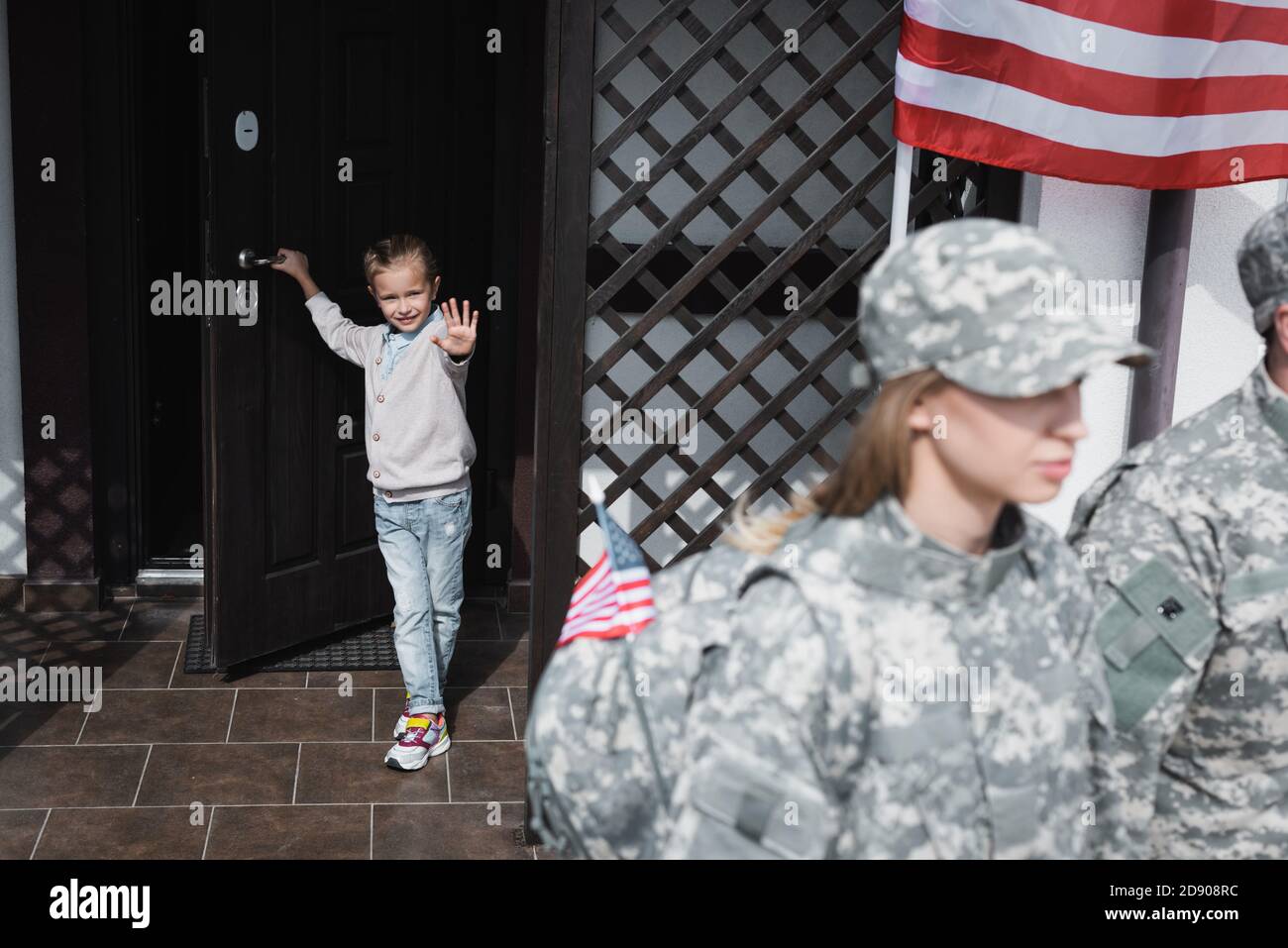 Smiling daughter with waving hand standing in doorway near american flag with blurred mother and father in military uniforms on foreground Stock Photo