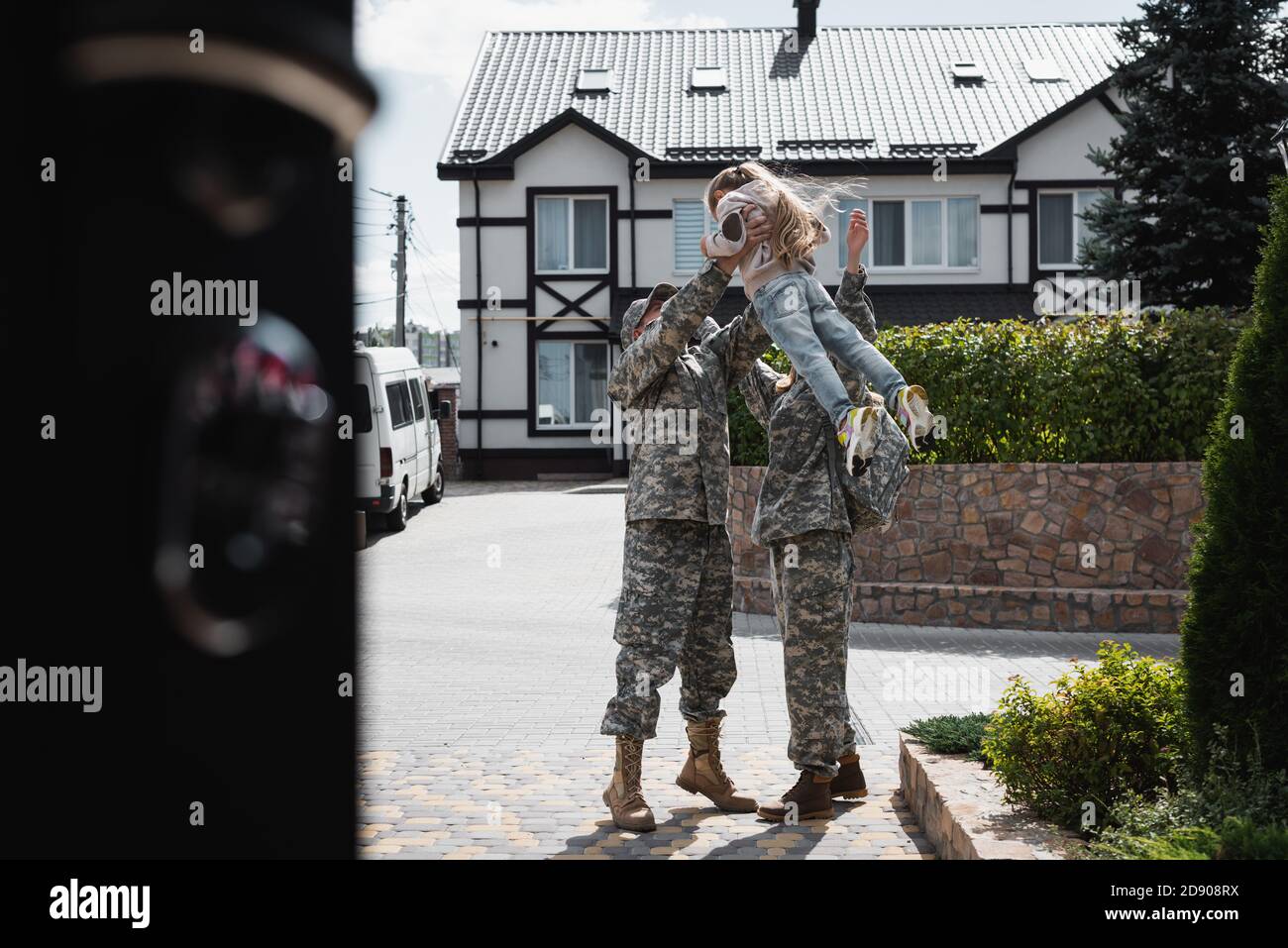 Father and mother in military uniforms lifting daughter in air, while standing on street on blurred foreground Stock Photo
