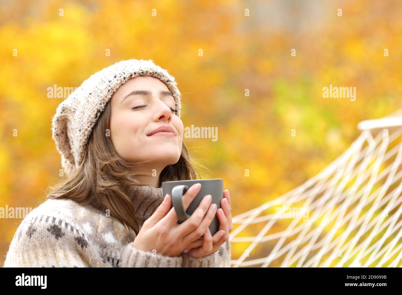Relaxed woman breaths fresh air holding coffee cup for breakfast sitting on a rope hammock in autumn in a forest Stock Photo