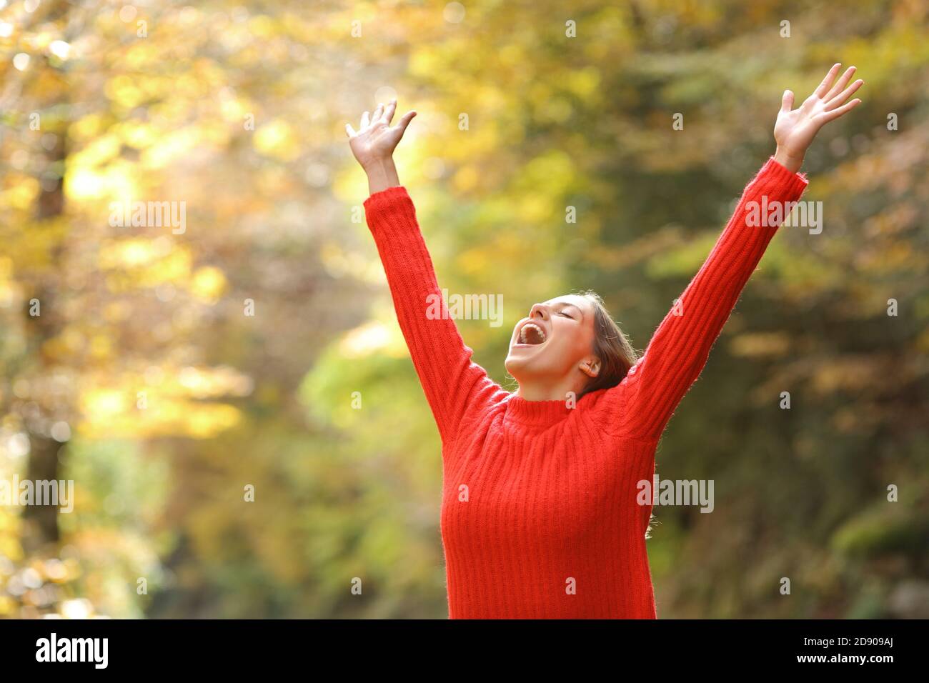Excited woman wearing red sweater celebrating success raising arms in a forest or park in autumn season Stock Photo