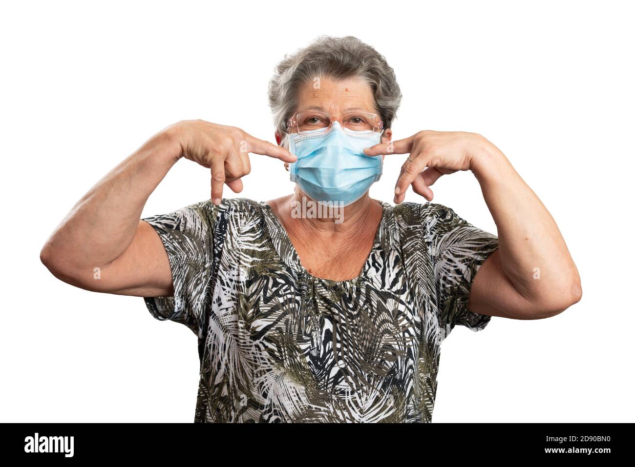 Old female person wearing glasses pointing index fingers at disposable sars covid19 virus medical or surgical protection mask as prevent pandemic conc Stock Photo