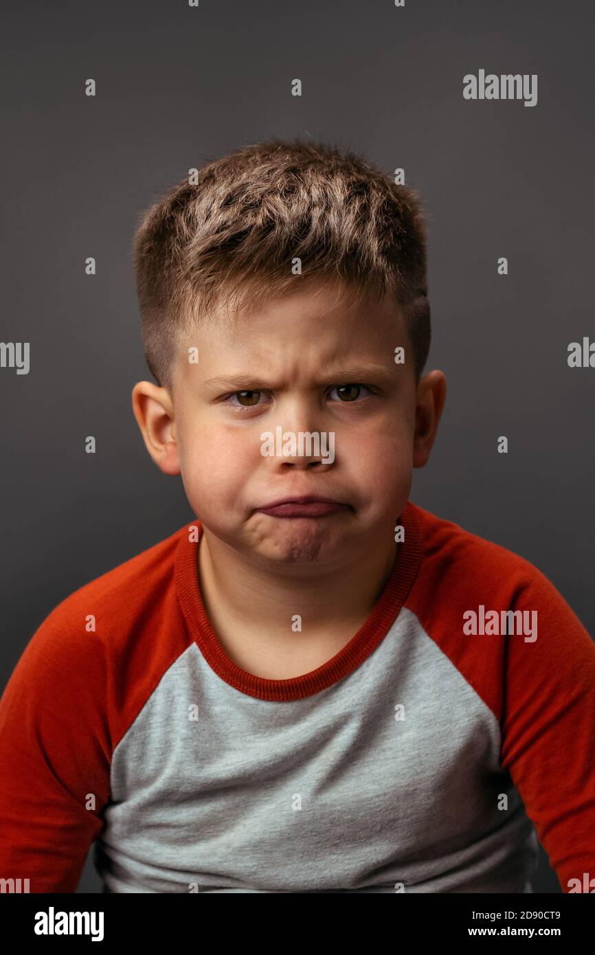 Little boy is naughty and grimaces with displeasure. Disgruntled child shows his emotions. Studio portrait on gray background Stock Photo