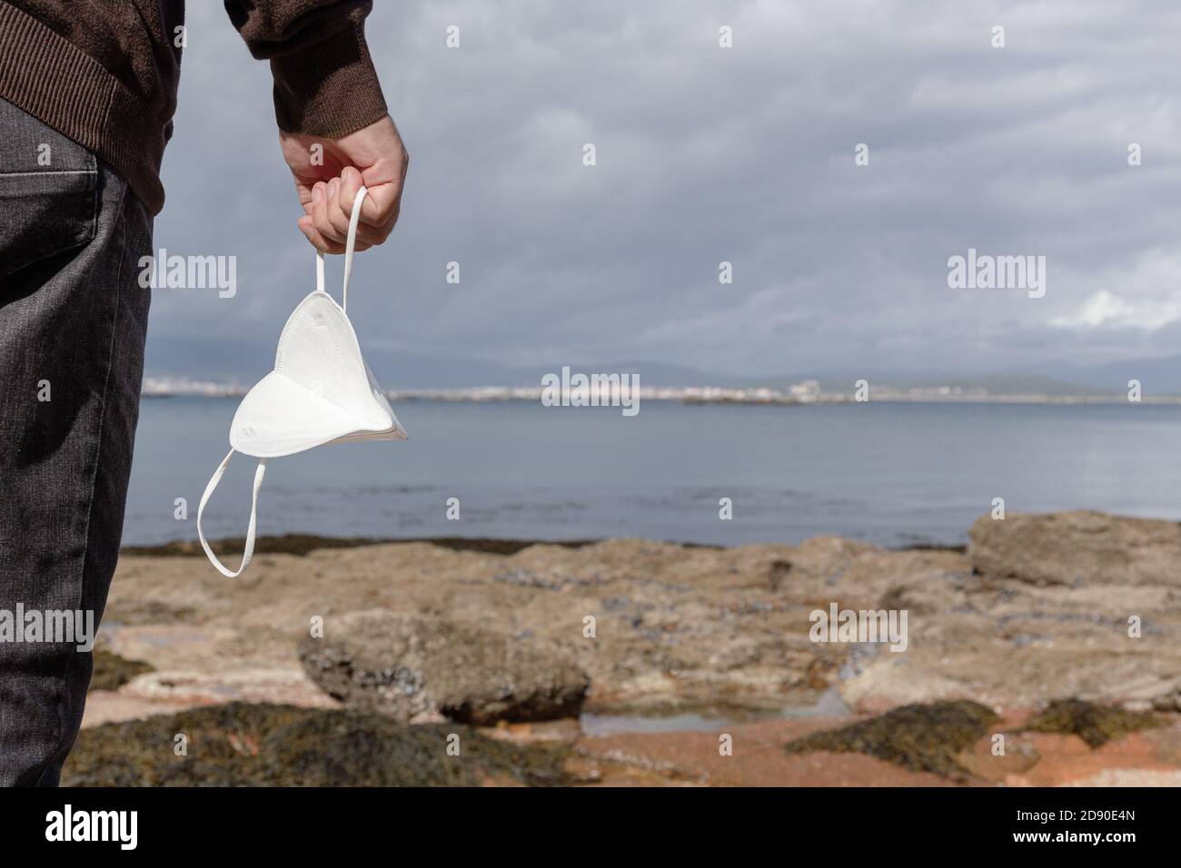 Male hand holding a face mask on a wild beach during a sunny day. Outdoor Freedom during Covid-19 pandemic concept. Copy space Stock Photo