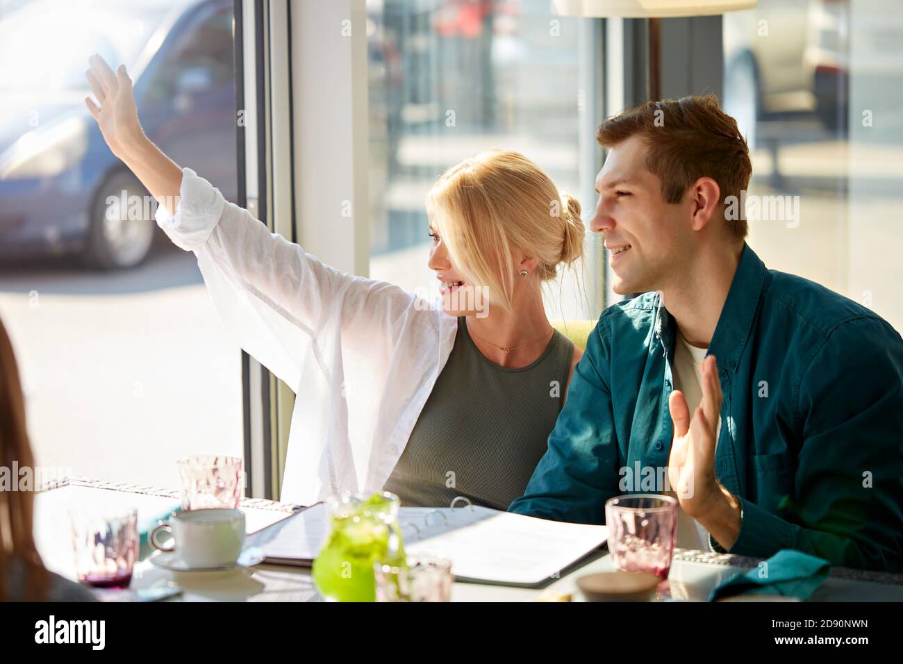 young beautiful couple in cafe have seen someone through window, they were waiting for friends to spend time together in restaurant. they show hello gesture at side Stock Photo