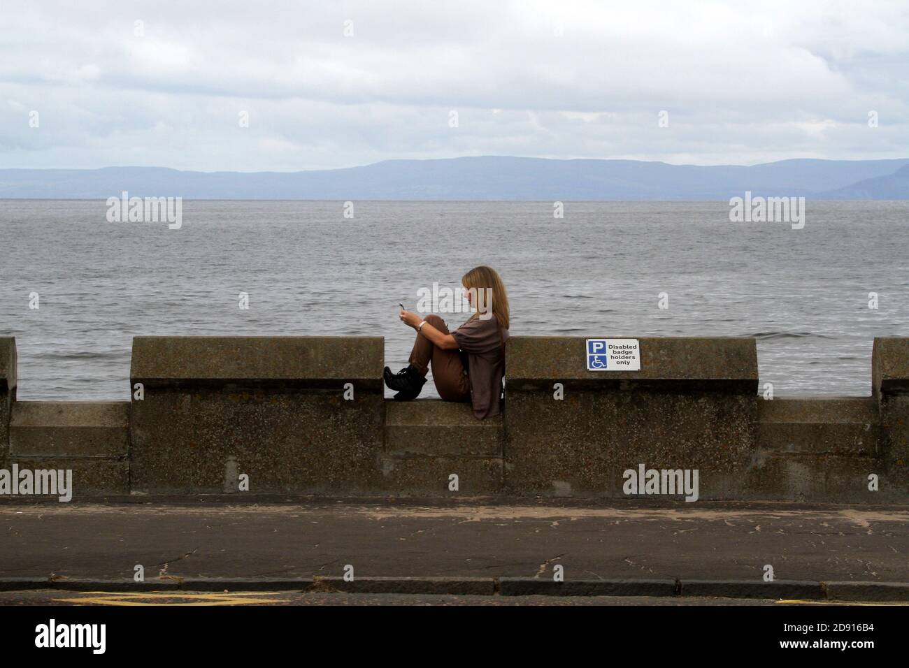 Ayr, Ayrshire, Scotland, UK  Young Woman sat on sea wall texting using her mobile phone Stock Photo