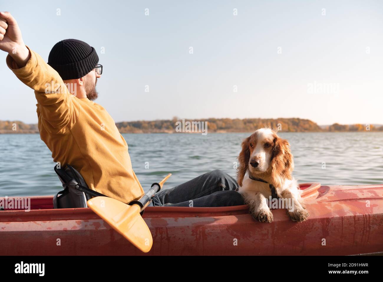 Kayaking with dogs: man stretches sitting in a row boat on the lake next to his spaniel. Active rest and adventures with pets, riding a canoe with dog Stock Photo