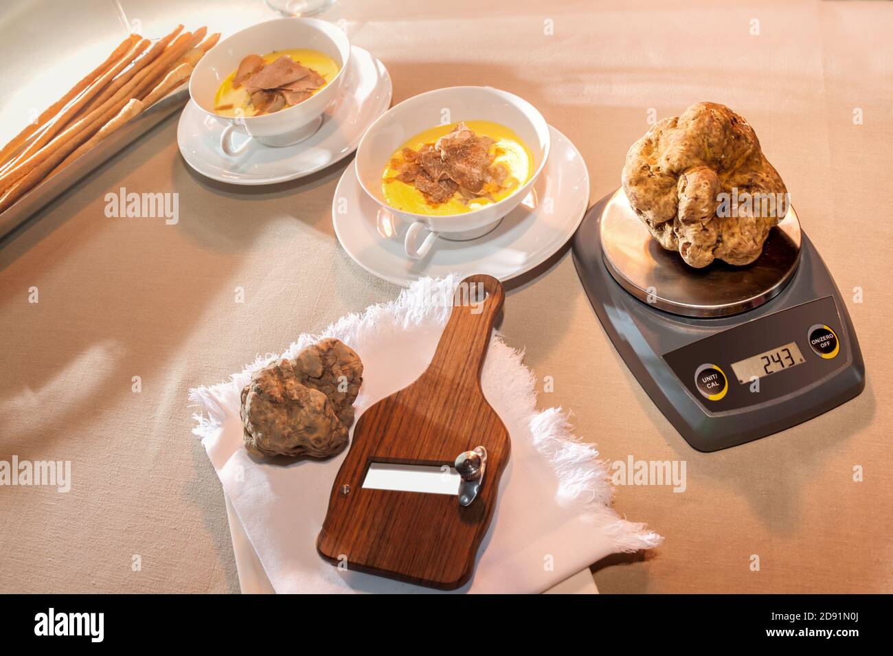 scale weighing a large beautiful white truffle near wooden slicer in the background two cups with cheese fondue and sliced truffles, all on the table Stock Photo