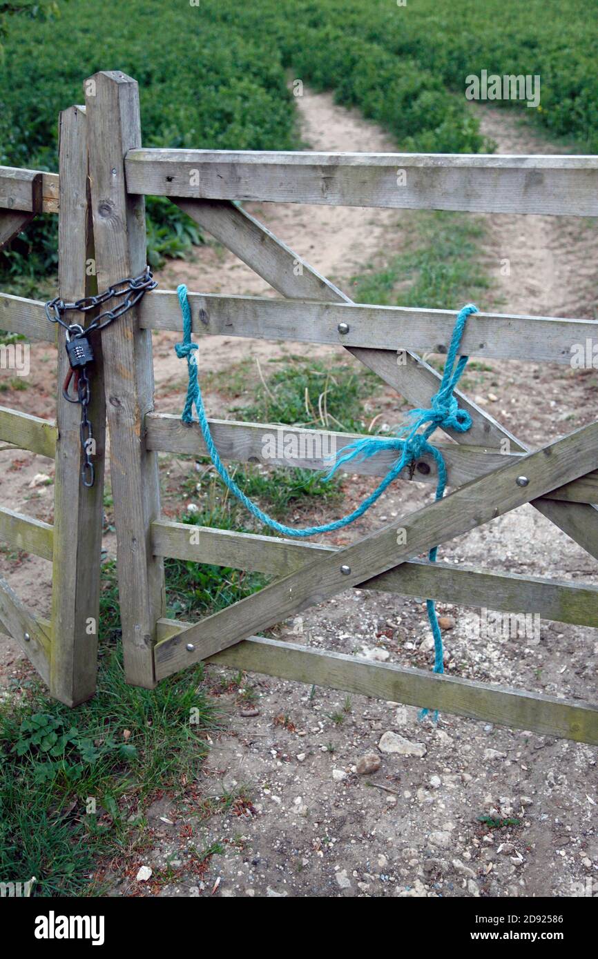 A 5-bar wooden gate at the entrance to a field, barely standing and held together by a chain and combination lock. A blue rope is attached to one gate. Stock Photo