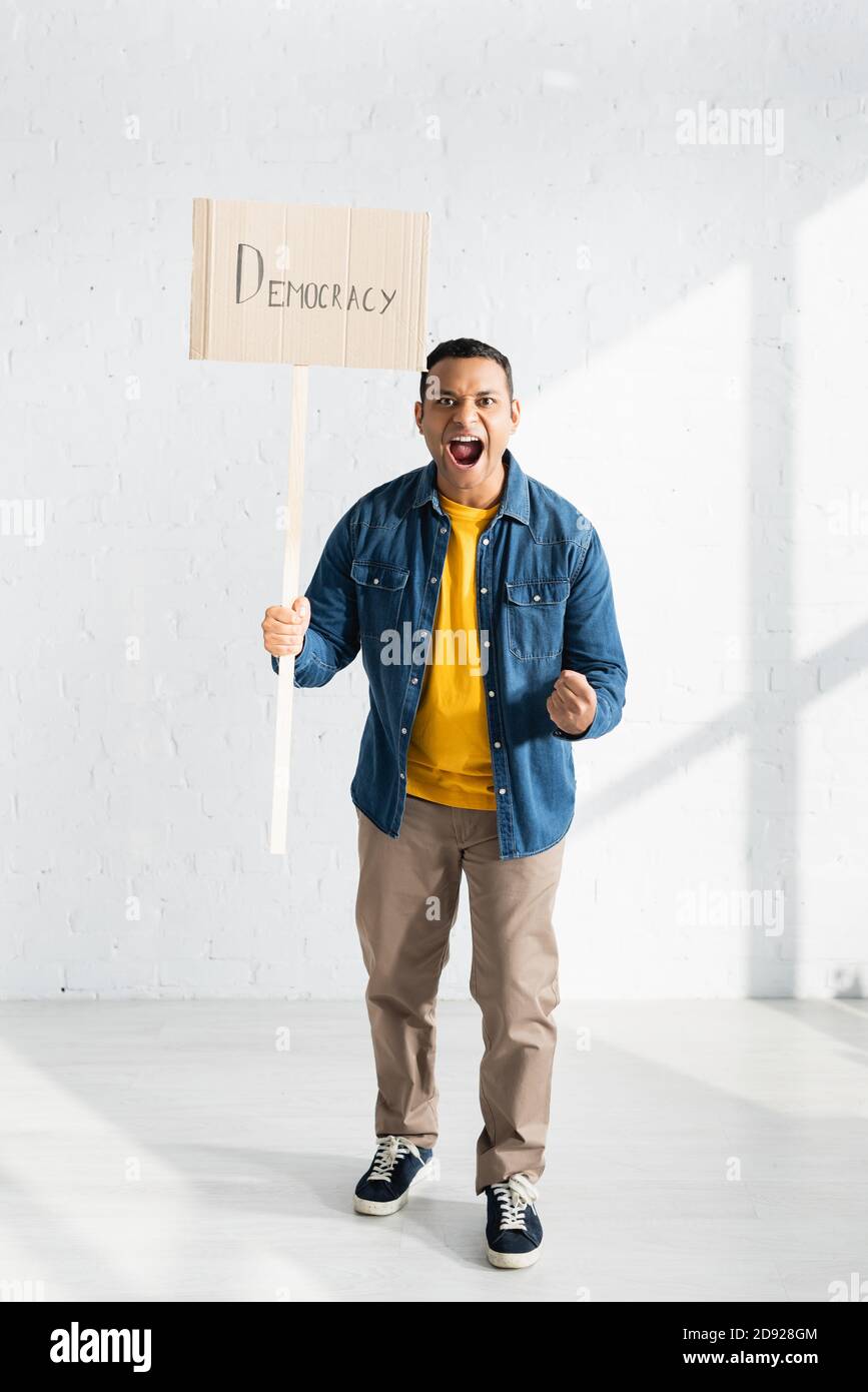 aggressive indian man screaming while holding placard with democracy lettering against white brick wall Stock Photo