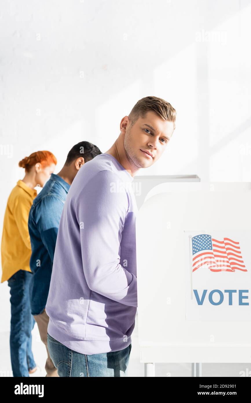 man looking at camera near polling booth with american flag and vote inscription, and multicultural electors on blurred background Stock Photo