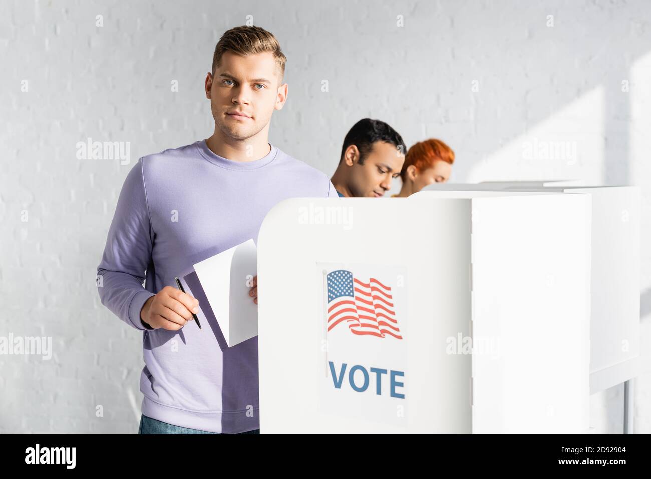 man looking at camera while holding ballot near polling booth with american flag and vote lettering on blurred background Stock Photo