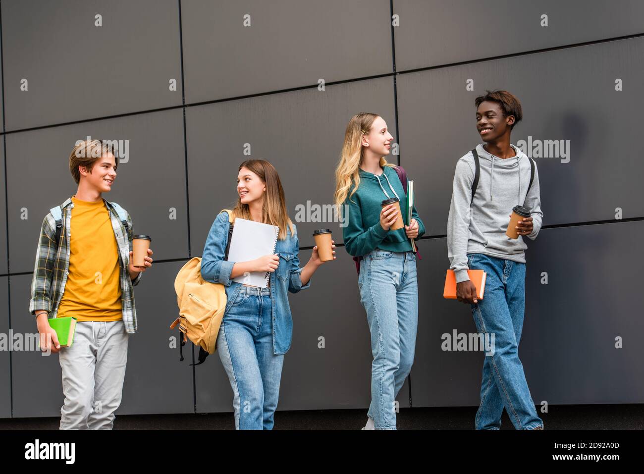 Positive multiethnic teenagers with backpacks and coffee to go walking near building Stock Photo