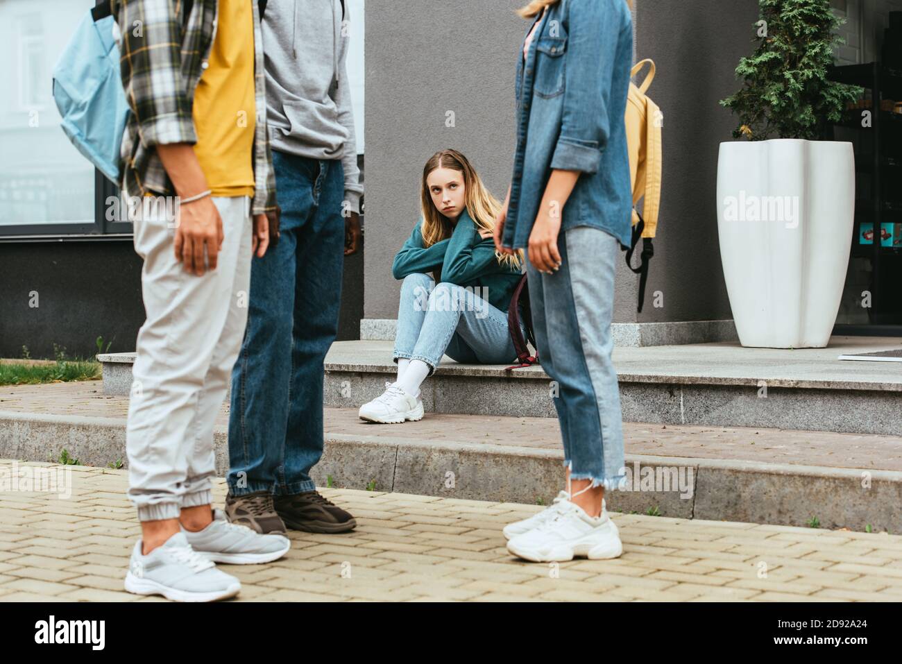 Selective focus of sad girl looking at multiethnic teenagers with backpacks outdoors Stock Photo