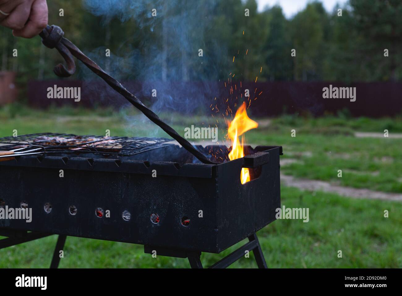 The meat is grilled. A man stirring coals with a hot poker. Sparks fly from the fire in the grill. Outdoor cooking. Meeting with friends, picnic, week Stock Photo