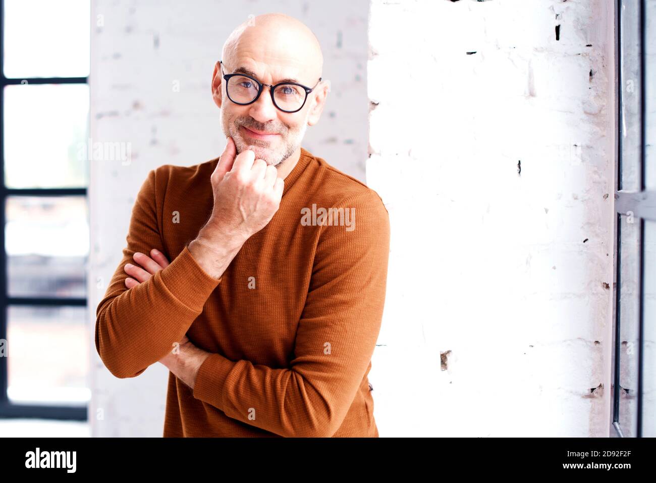 Portrait of curious man with beard and eyewear standing at the window. Hand on chin. Stock Photo