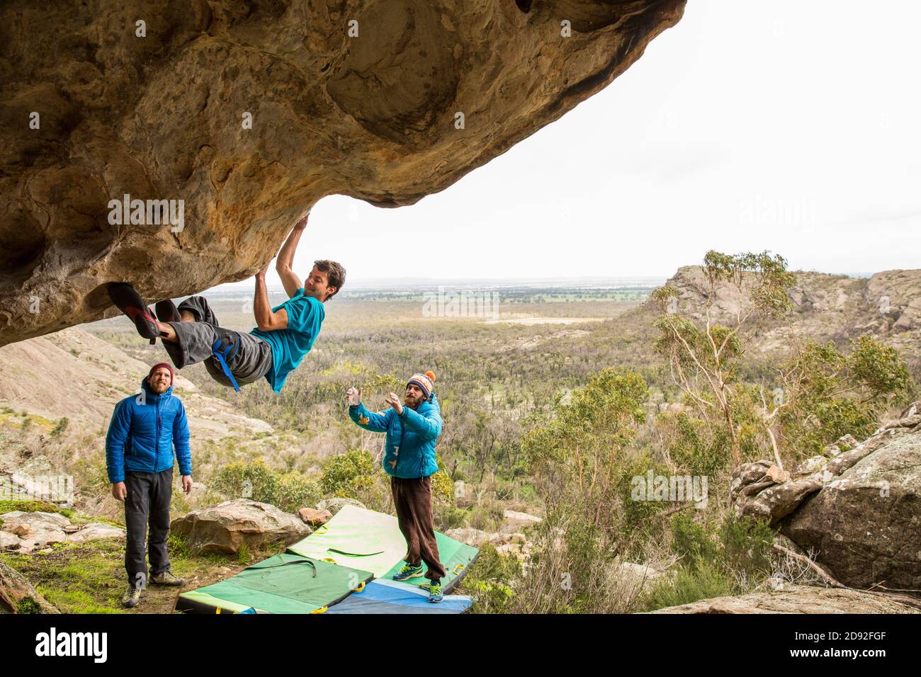 Athletic male climbs outside on a boulder with men spotting Stock Photo