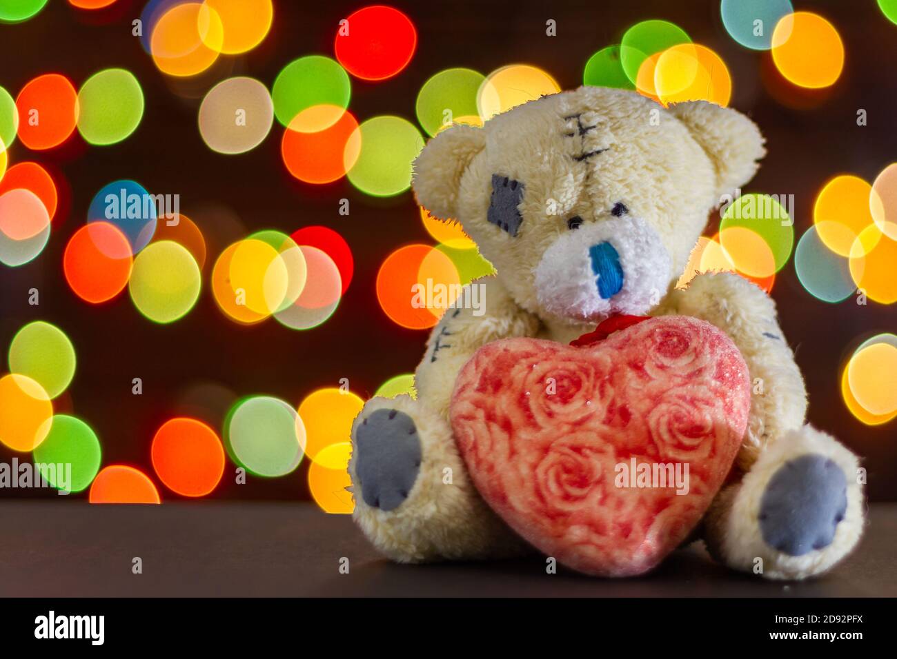 Soft toy bear with heart on the background of bokeh of colorful lights of garlands. Copy space for text, selective focus Stock Photo