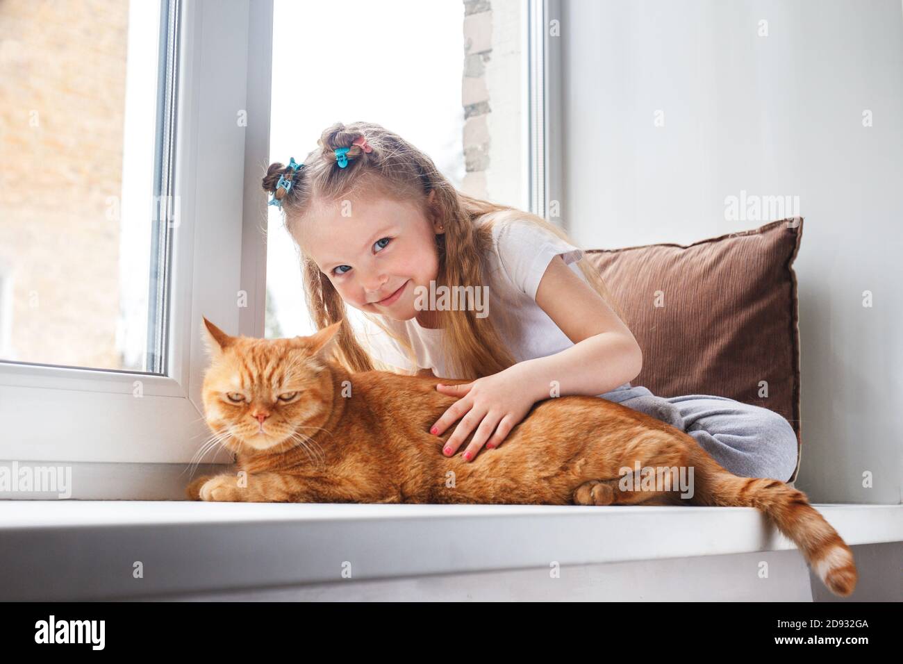 Cute child embraces with tenderness and love a red cat on window Stock Photo